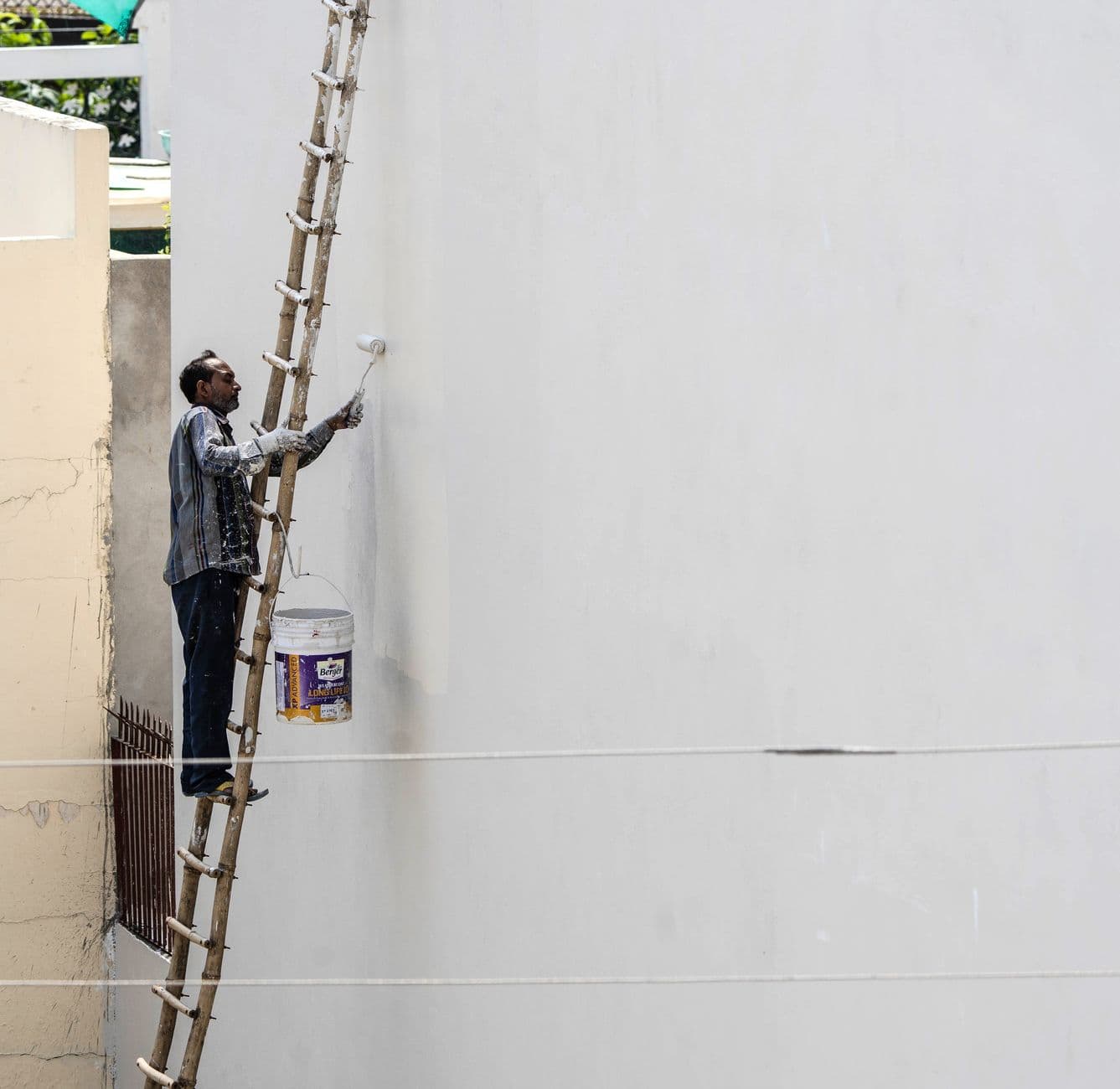 A man on a ladder painting a building's exterior wall with a roller brush outdoors.